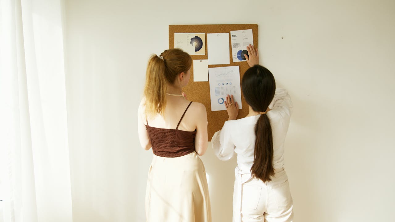 Two women analyzing charts and graphs on a corkboard. Business teamwork concept.